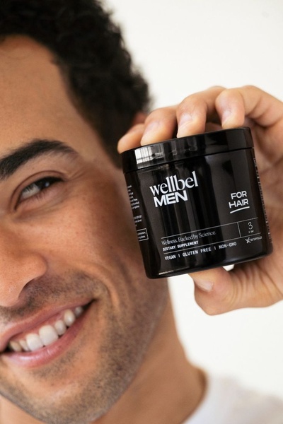 A close-up photograph featuring a smiling man holding a jar of Wellbel Men supplement, emphasizing personal health and brand clarity.